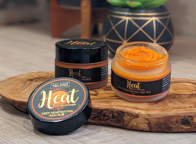 The Blend Heat Pain Balm 2 jars one with lid open on wooden base with plant in background